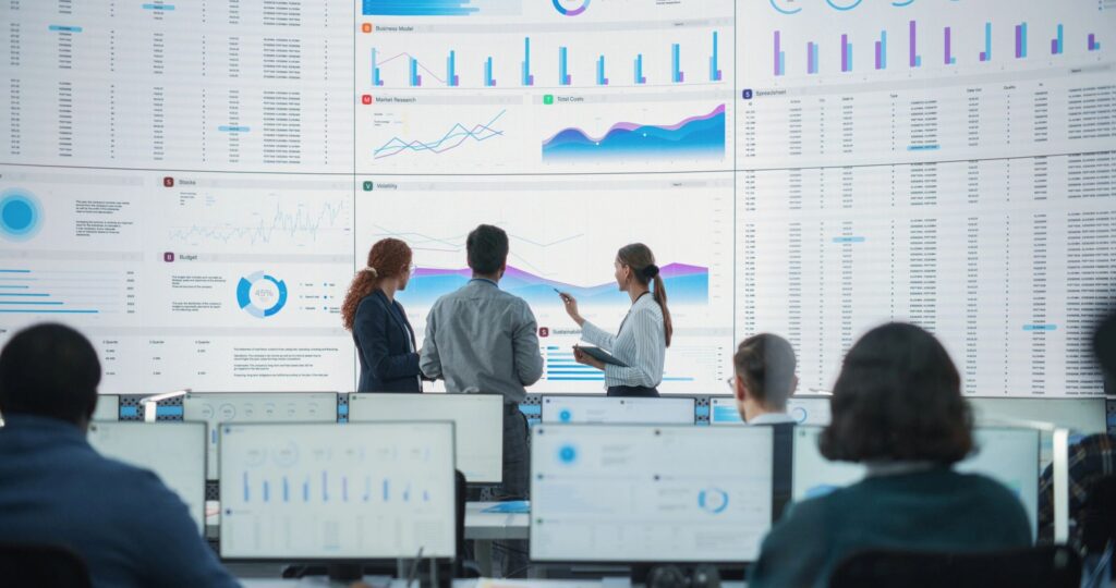 Wide Back Shot of Diverse Team of Data Scientists Discussing Graphs And Reports On Big Digital Screen In Monitoring Office. Multiethnic Employees Working On Desktop Computers For Consulting Company.